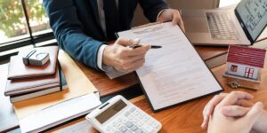 Person pointing at a purchase agreement on a clipboard, with a calculator and a small house model on the desk.
